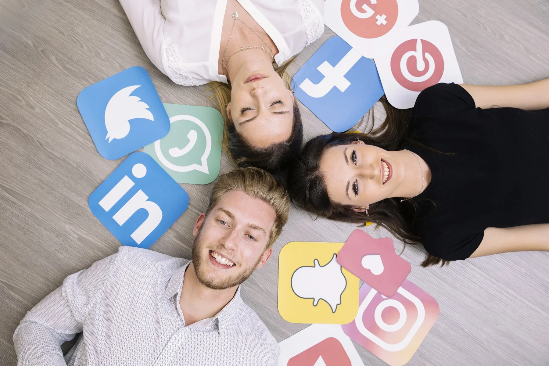 High-angle view of three smiling young adults lying on a wooden floor surrounded by social media icons like LinkedIn, WhatsApp, Instagram, Snapchat, Facebook, and others, representing Multi-Channel Marketing strategies.